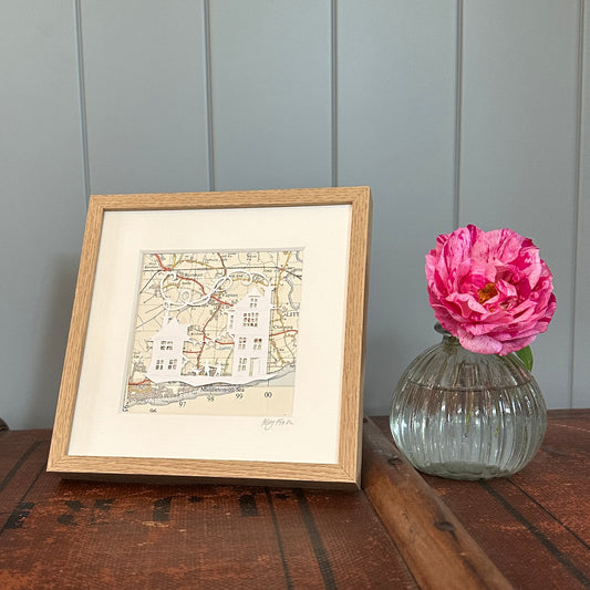 white papercut of two buildings with bunting and tow people and a dog overlaid onto an OS Map extract in brown wood effect frame next to flower for scale