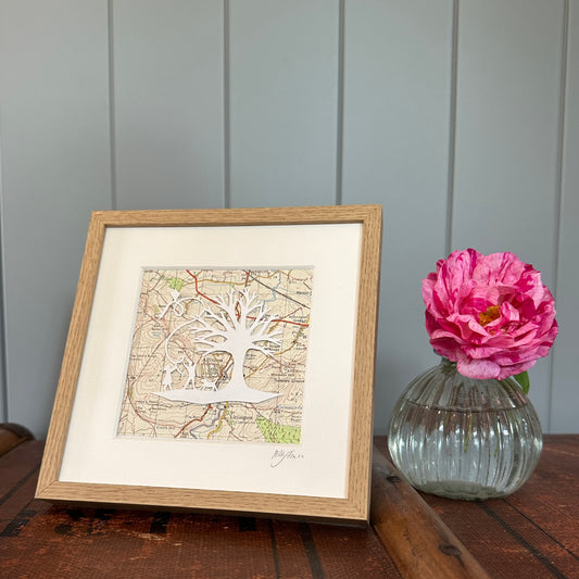 Papercut showing kite flying next to a tree overlaid onto an OS map extract in wood effect frame next to a flower for scale