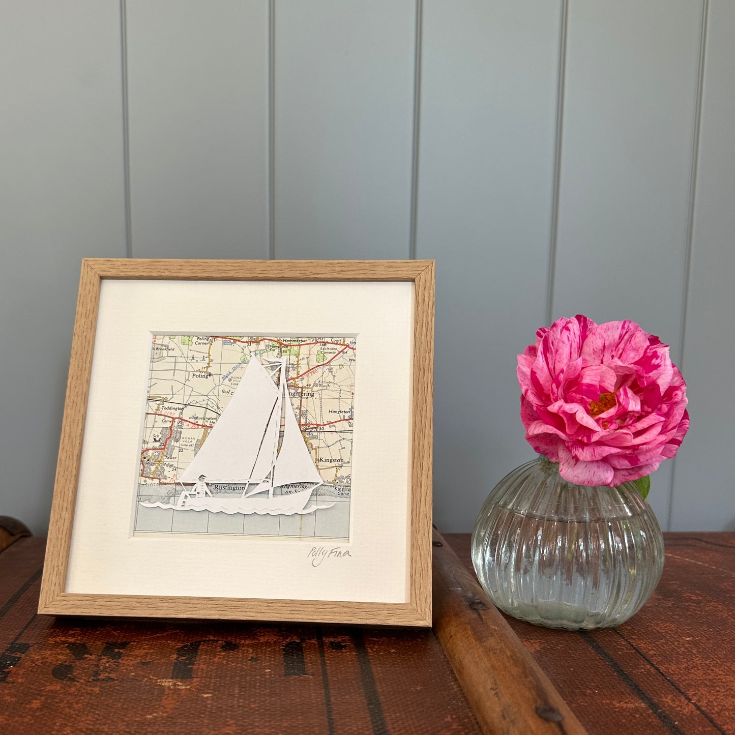 Papercut of sailing dinghy over an OS map extract in wood effect frame next to flower for scale