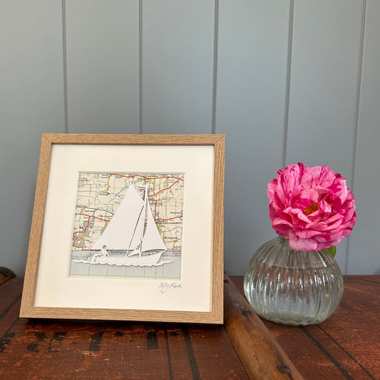 Papercut of sailing dinghy over an OS map extract in wood effect frame next to flower for scale