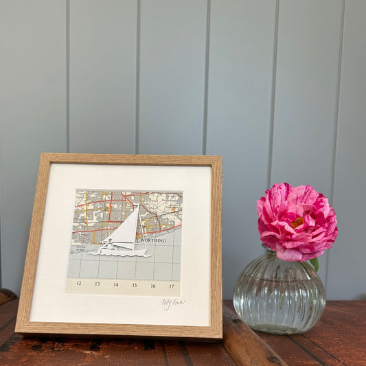 papercut sailing boat over OS map extract in wood effect frame next to flower for scale